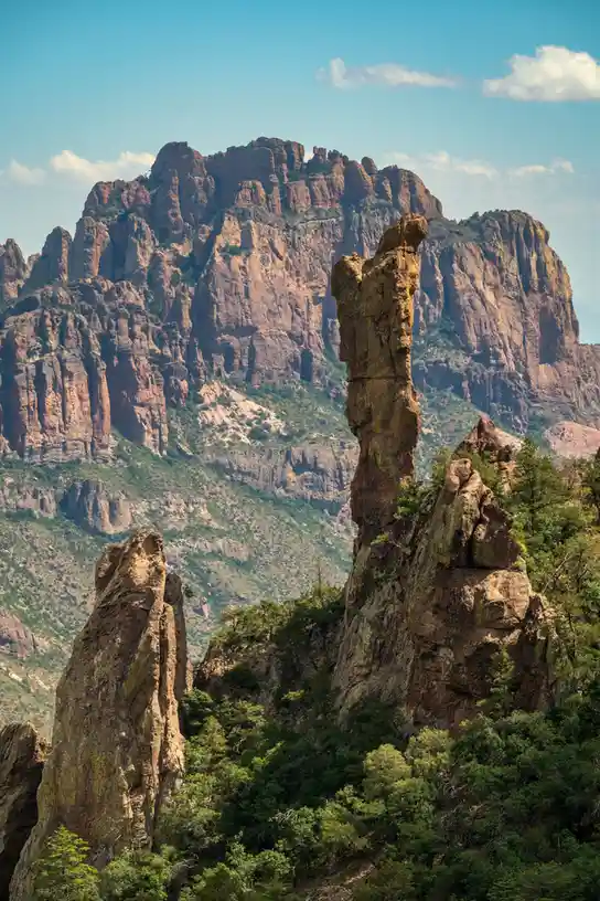 A scenic photograph of a rugged, red rock canyon with towering rock formations and green trees under a blue sky with clouds…