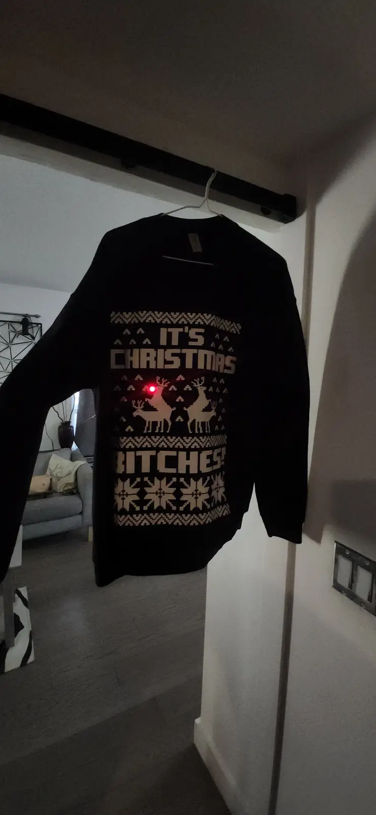 A black Christmas sweater hangs on a hook, featuring the text 'IT'S CHRISTMAS BITCHES!' in a festive pattern with reindeer…