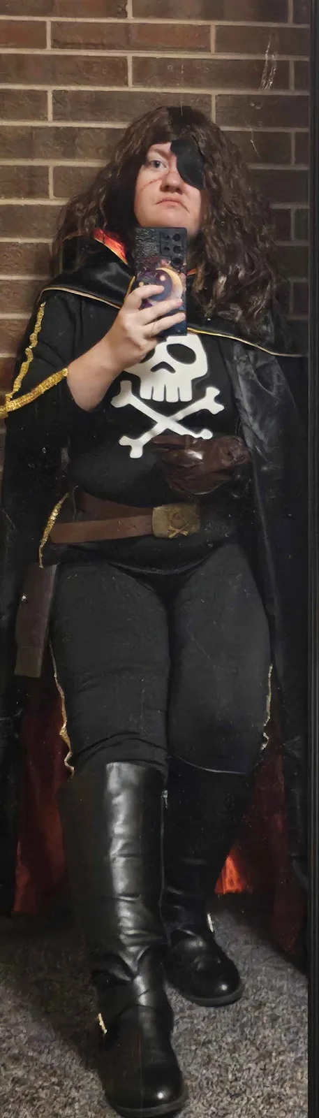 A person in a pirate costume with a black eye patch, skull and crossbones shirt, and black boots takes a mirror selfie while…