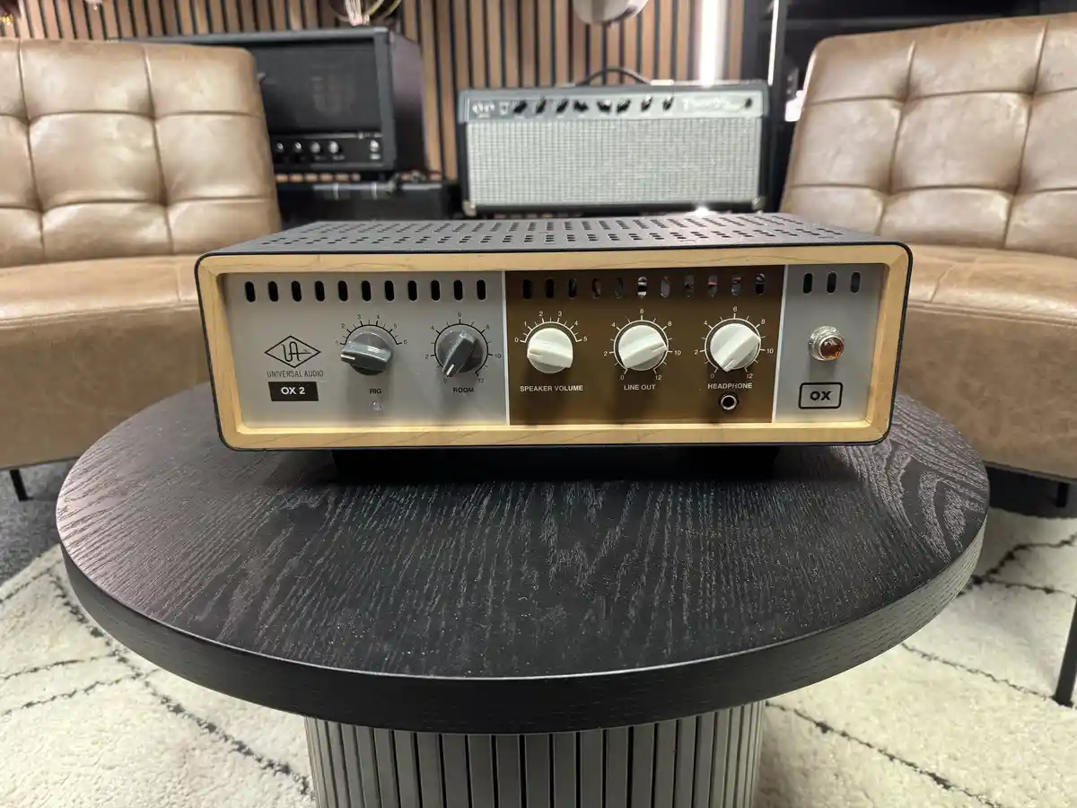 A Universal Audio OX 2 audio interface sits on a dark wooden table, with two leather couches and vintage guitar amplifiers…