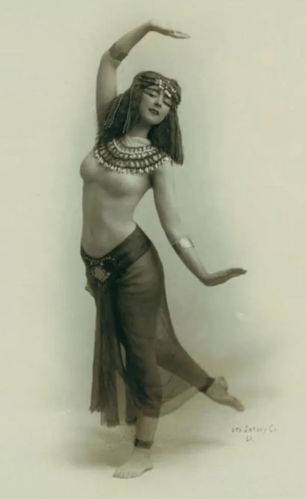 A vintage sepia-toned photograph shows a woman in an ornate, flowing costume with a headpiece and beaded necklace, striking…