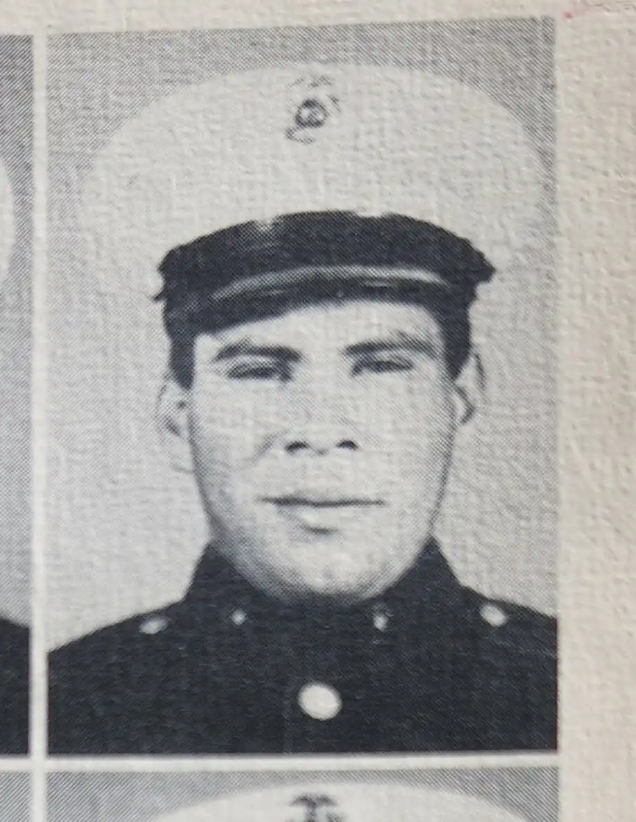A grainy black-and-white photo shows a young man in a military uniform and cap, looking directly at the camera with a…