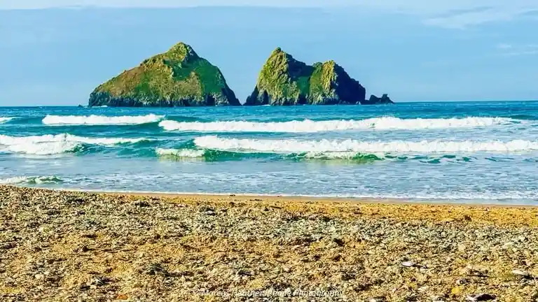 A scenic beach photo shows turquoise waves rolling onto a pebbly shore with two large, green-covered sea stacks in the…