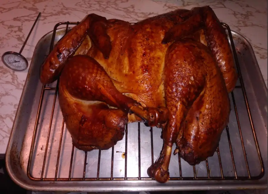 A roasted turkey sits on a metal rack inside a baking tray, its skin golden-brown and crispy. A meat thermometer rests on…