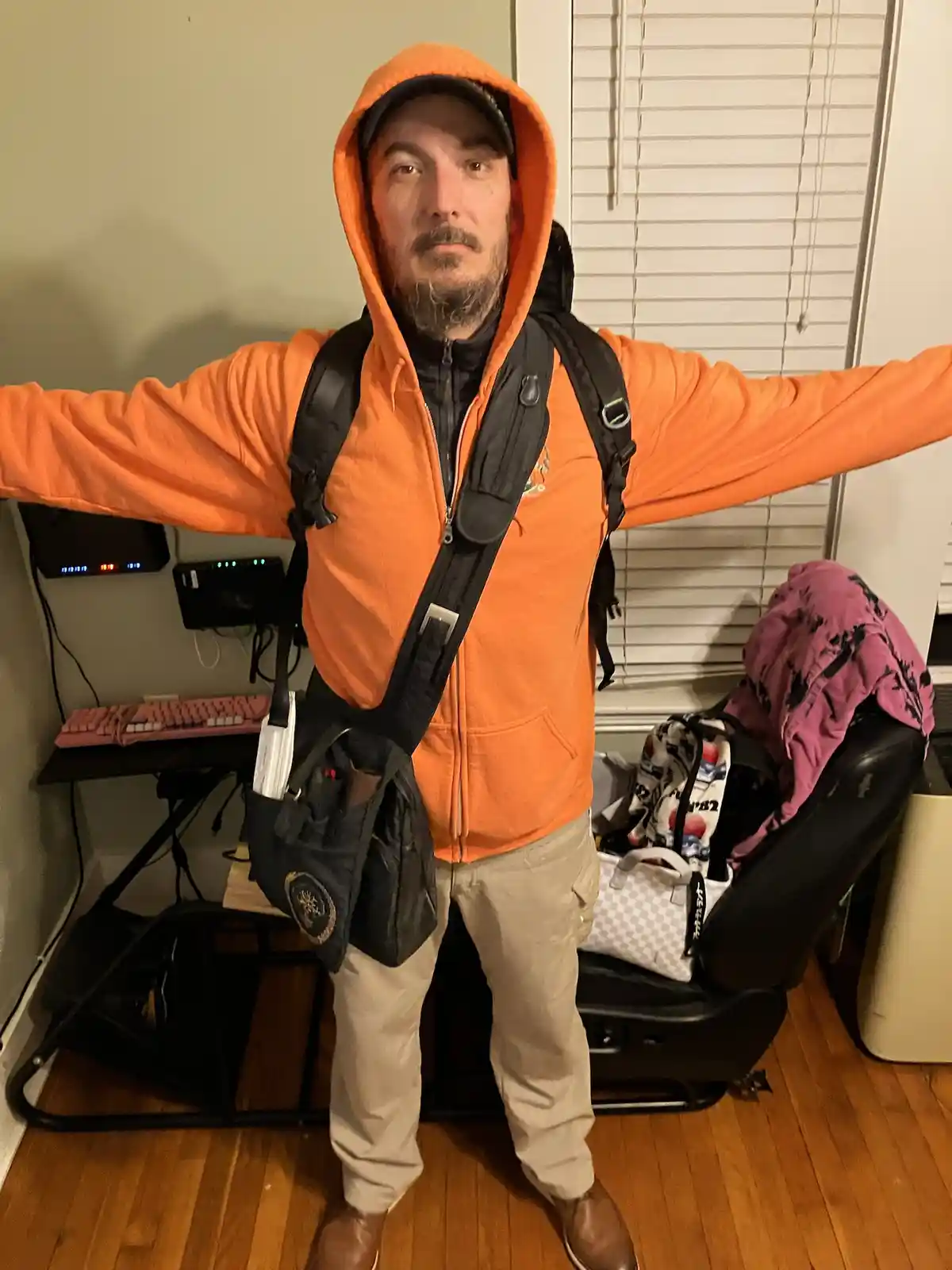 A man in an orange hooded jacket and beige pants stands indoors with arms spread wide, wearing a black tactical-style…