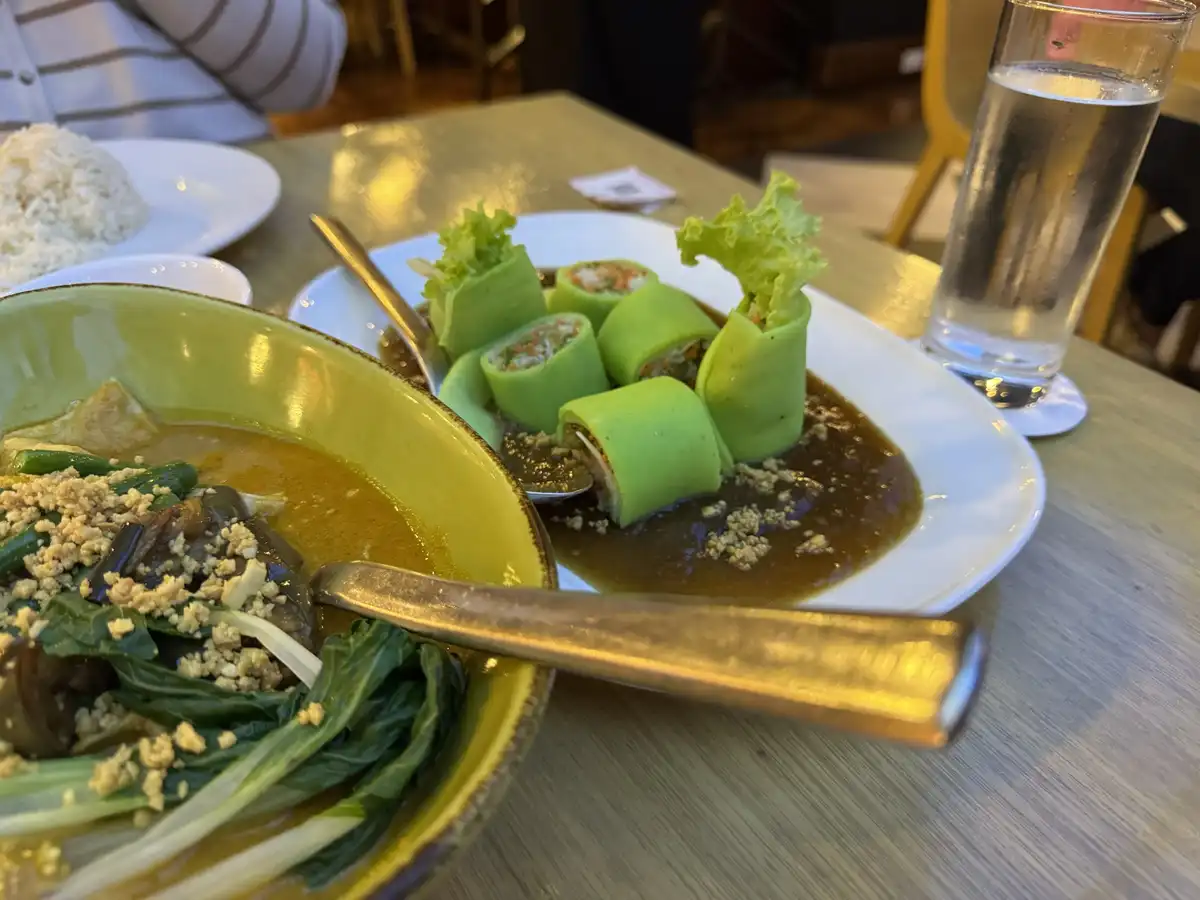 A close-up photo of a restaurant table with two dishes: a yellow bowl of green curry with eggplant and greens, and a white…
