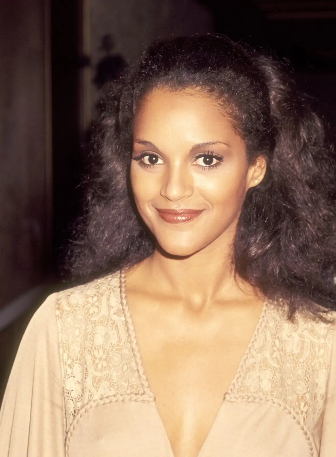 A vintage photo of a woman with voluminous, dark, curly hair styled in a high ponytail with side bangs, wearing a beige…