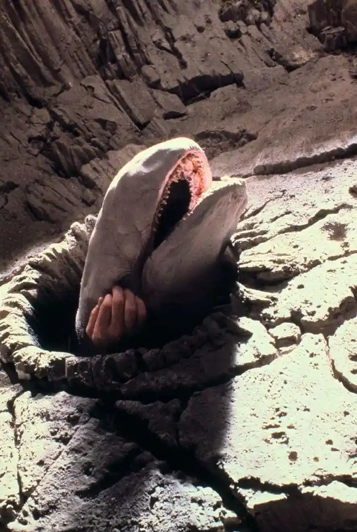 A person's hand is visible gripping the edge of a hole in cracked earth, with a large, monstrous-looking creature's head…