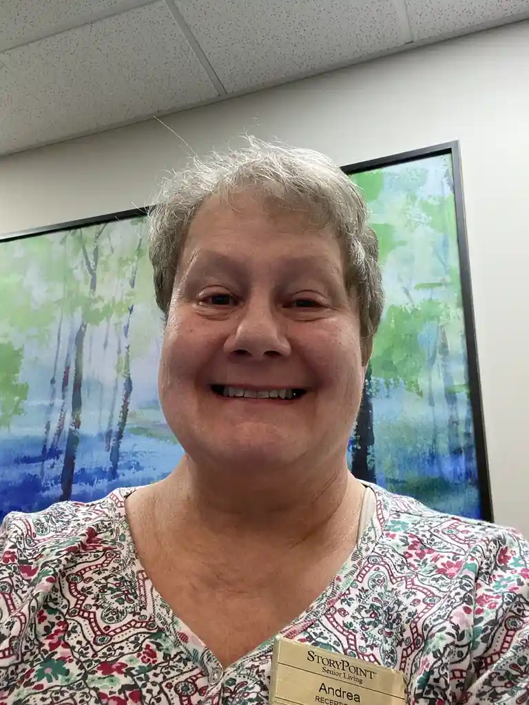 A smiling woman with short gray hair wears a name tag that reads 'STORYPOINT Senior Living' and 'Andrea RECEPT'. She is in…