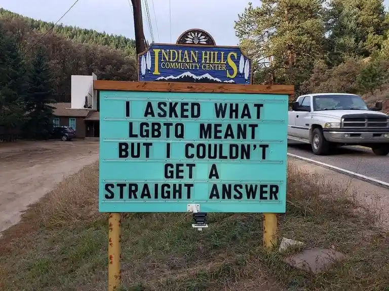 A turquoise outdoor sign with black lettering reads 'I ASKED WHAT LGBTQ MEANT BUT I COULDN'T GET A STRAIGHT ANSWER', placed…