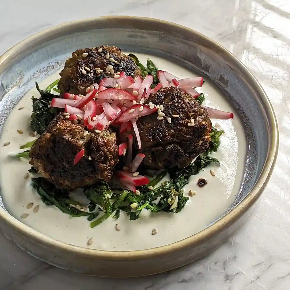 A close-up photo of a ceramic bowl containing three dark, seared meatballs resting on wilted greens and a creamy white…