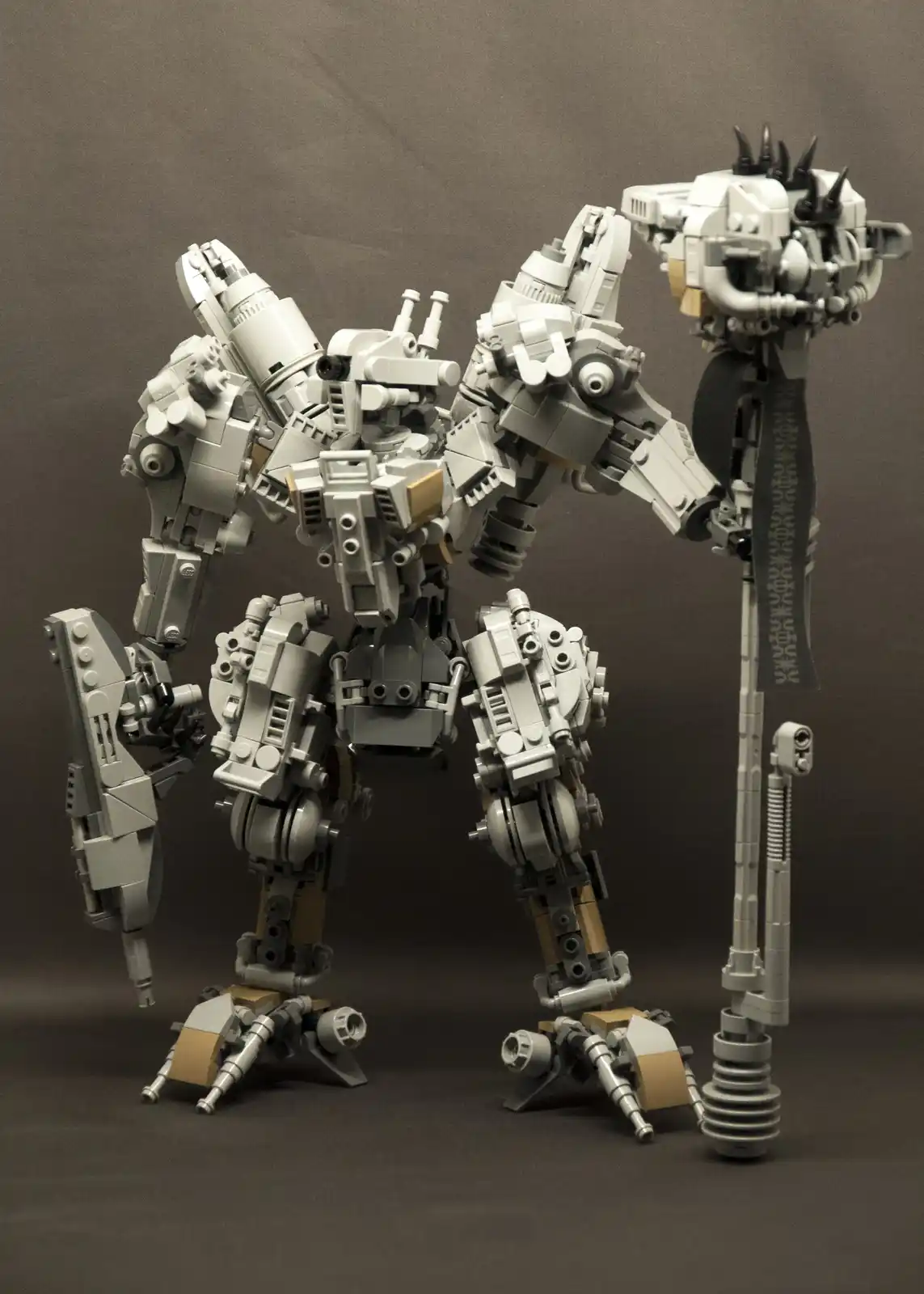 A highly detailed, grey and tan LEGO model of a bipedal mech or robot stands against a plain dark background, holding a long…