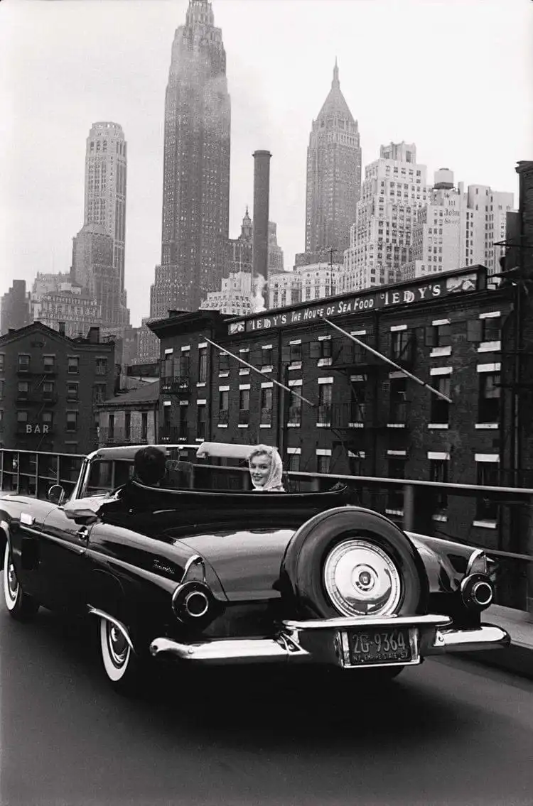 A black-and-white photograph shows a woman in a convertible car smiling at the camera while driving on a city street with a…