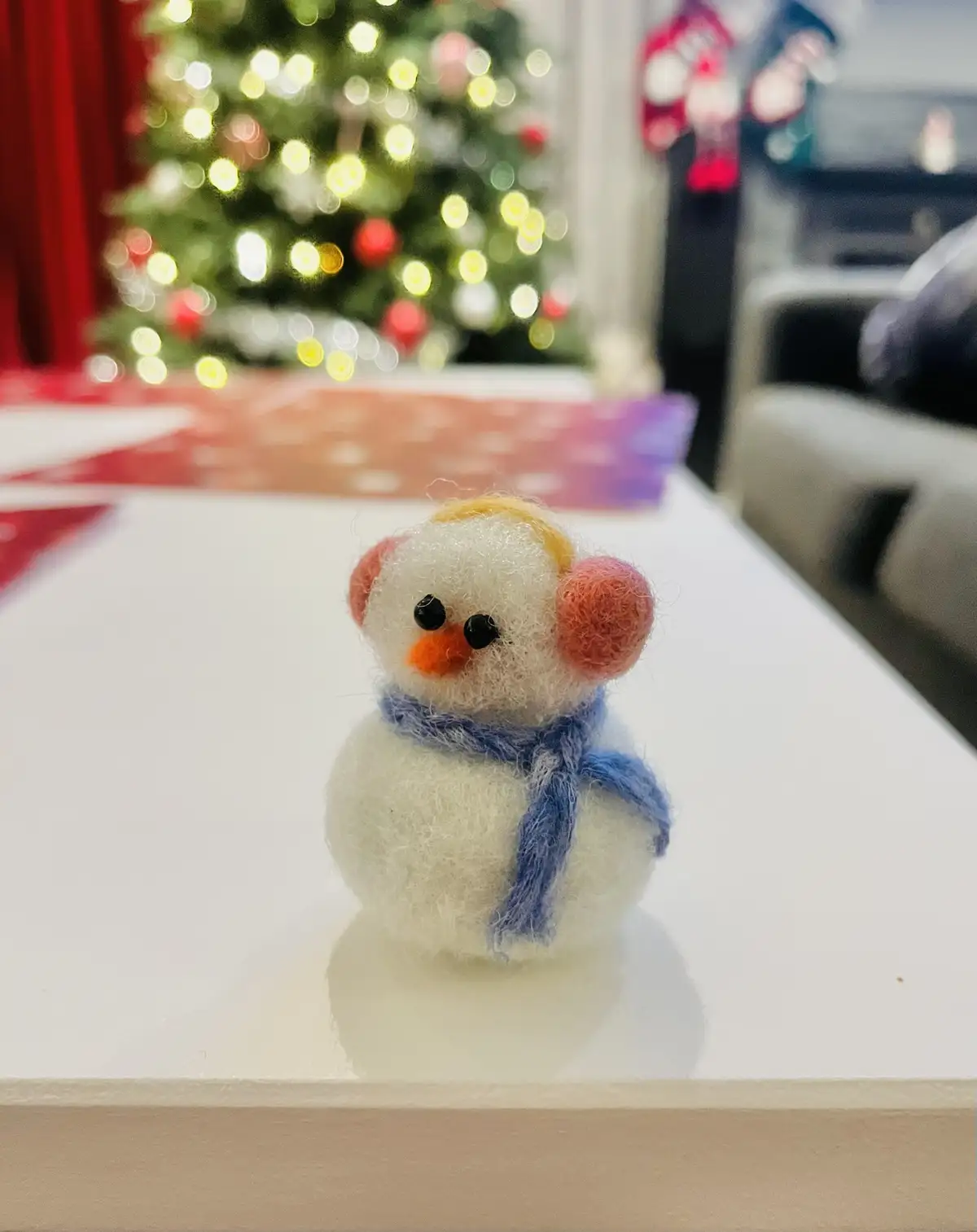 A small, handmade felt snowman figurine with pink earmuffs, a blue scarf, and an orange carrot nose sits on a white table…