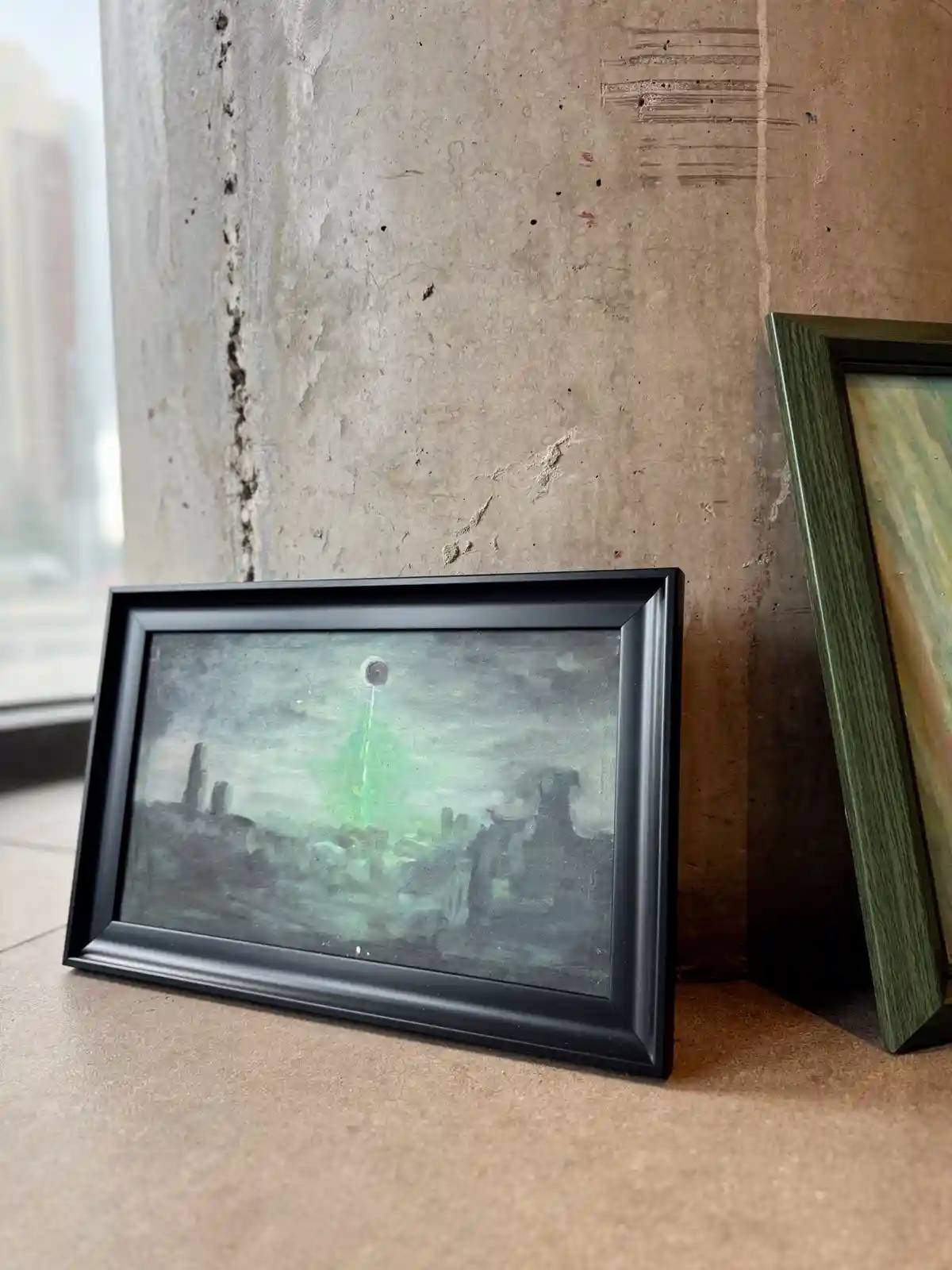A framed painting depicting a dark, gloomy cityscape with a green glowing beam shooting from a circular object in the sky…