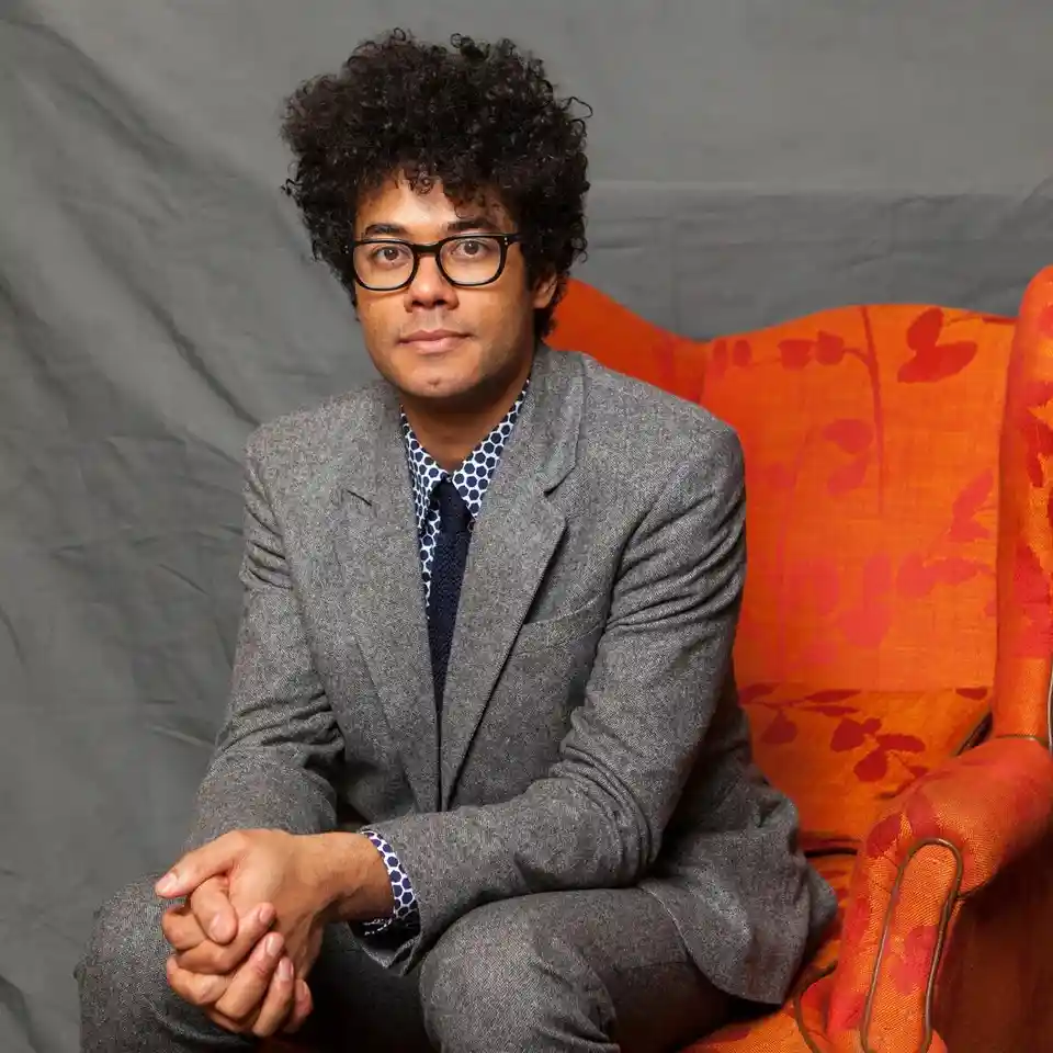 a man with curly hair and glasses sits in a gray suit on an orange floral armchair, looking directly at the camera with a…