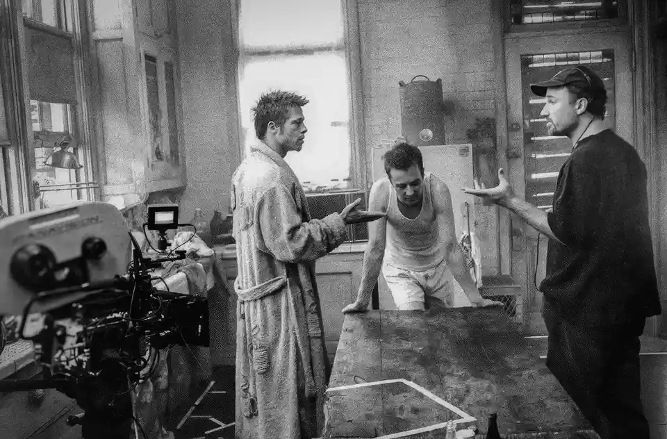 A black-and-white behind-the-scenes photo from a movie set shows three men in a gritty room. On the left, Brad Pitt in a…