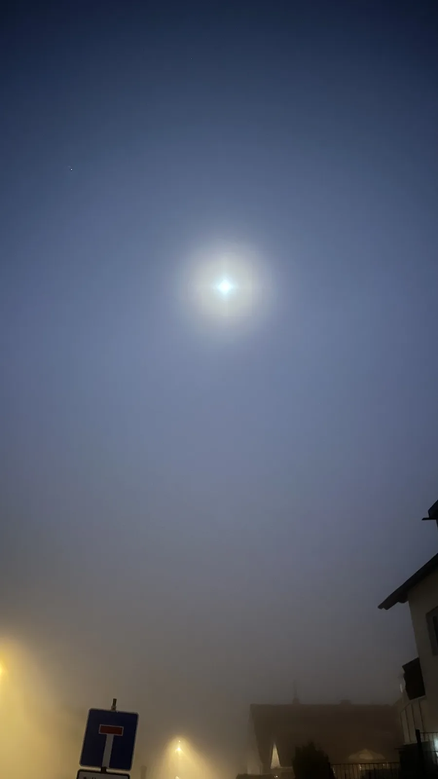 A foggy night sky with a bright moon and a faint star visible, with a blue road sign and building silhouettes at the bottom…