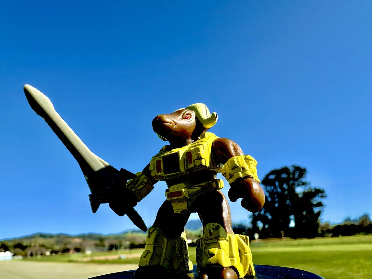 A toy figurine of a brown, bull-headed warrior in yellow armor, holding a large sword, stands outdoors against a clear blue…