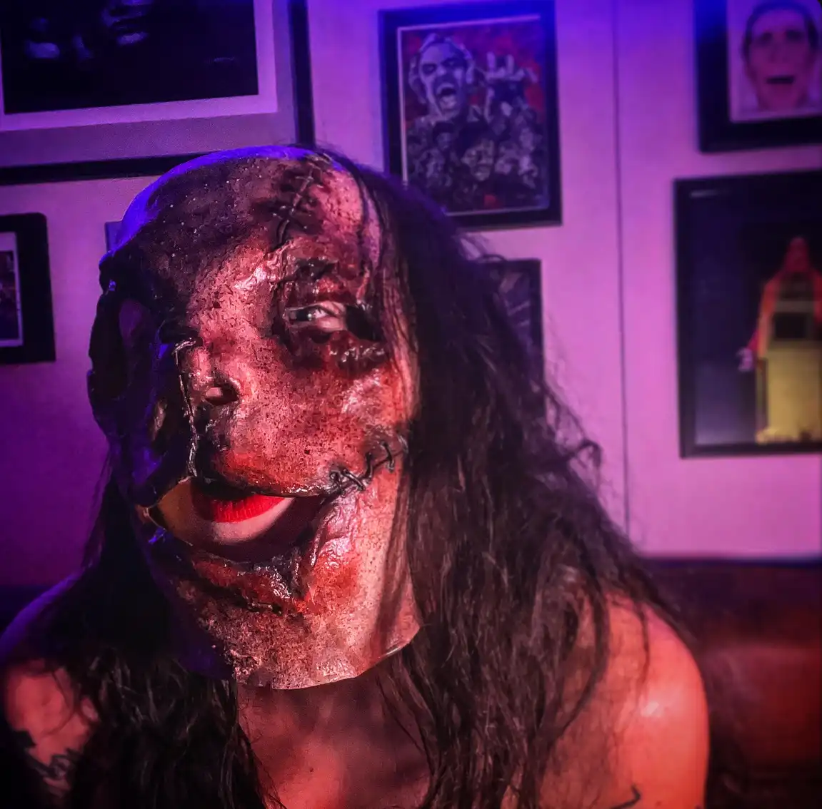 A person with long dark hair wears a grotesque, stitched-together horror mask with exposed wires and red lipstick, seated in…
