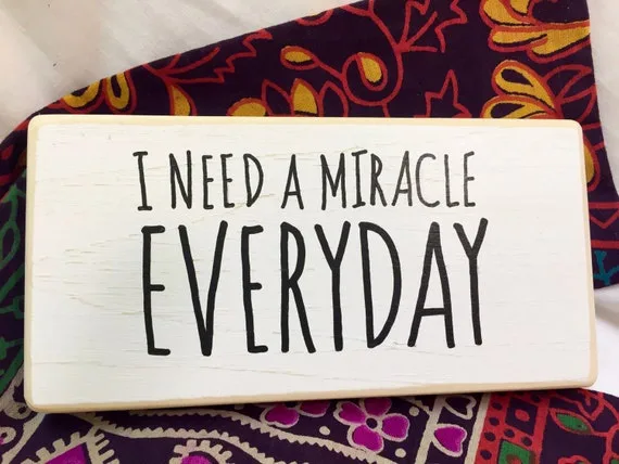 A white wooden sign with black handwritten text reading 'I NEED A MIRACLE EVERYDAY' rests on a colorful patterned fabric…
