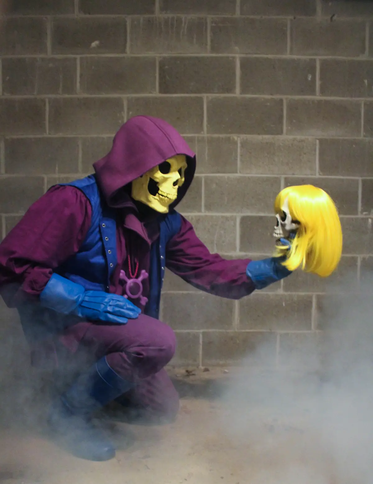 A person in a Skeletor costume, wearing a purple hooded outfit and blue gloves, kneels in front of a brick wall while…