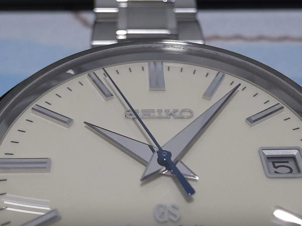 A close-up photo of a Seiko wristwatch with a cream-colored dial, silver rectangular hour markers, and a blue second hand…