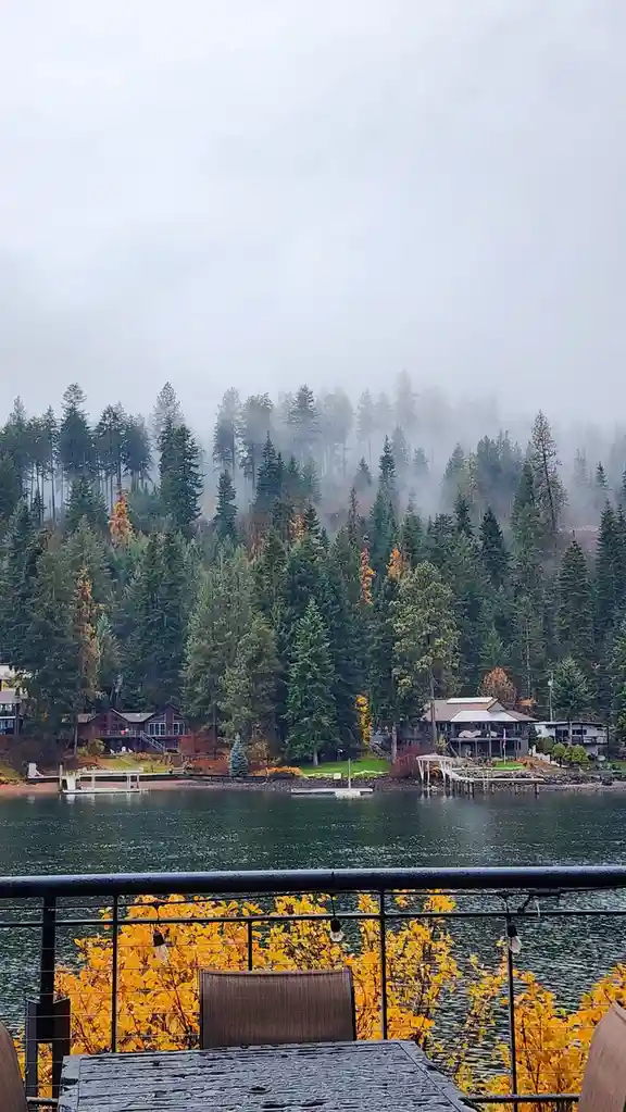 A rainy, foggy lakeside view from a balcony with a wet table and chair, overlooking houses and evergreen trees with some…