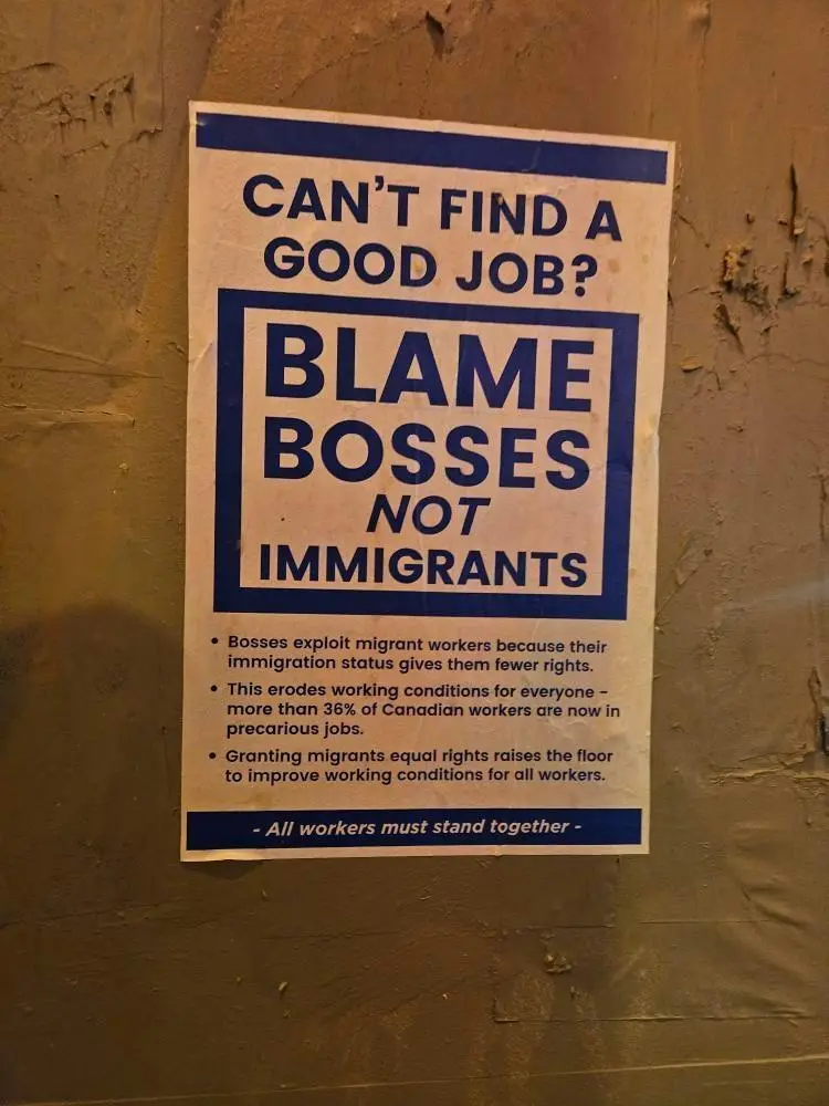 A poster on a textured wall reads 'CAN'T FIND A GOOD JOB? BLAME BOSSSES NOT IMMIGRANTS' in large blue text, with smaller…