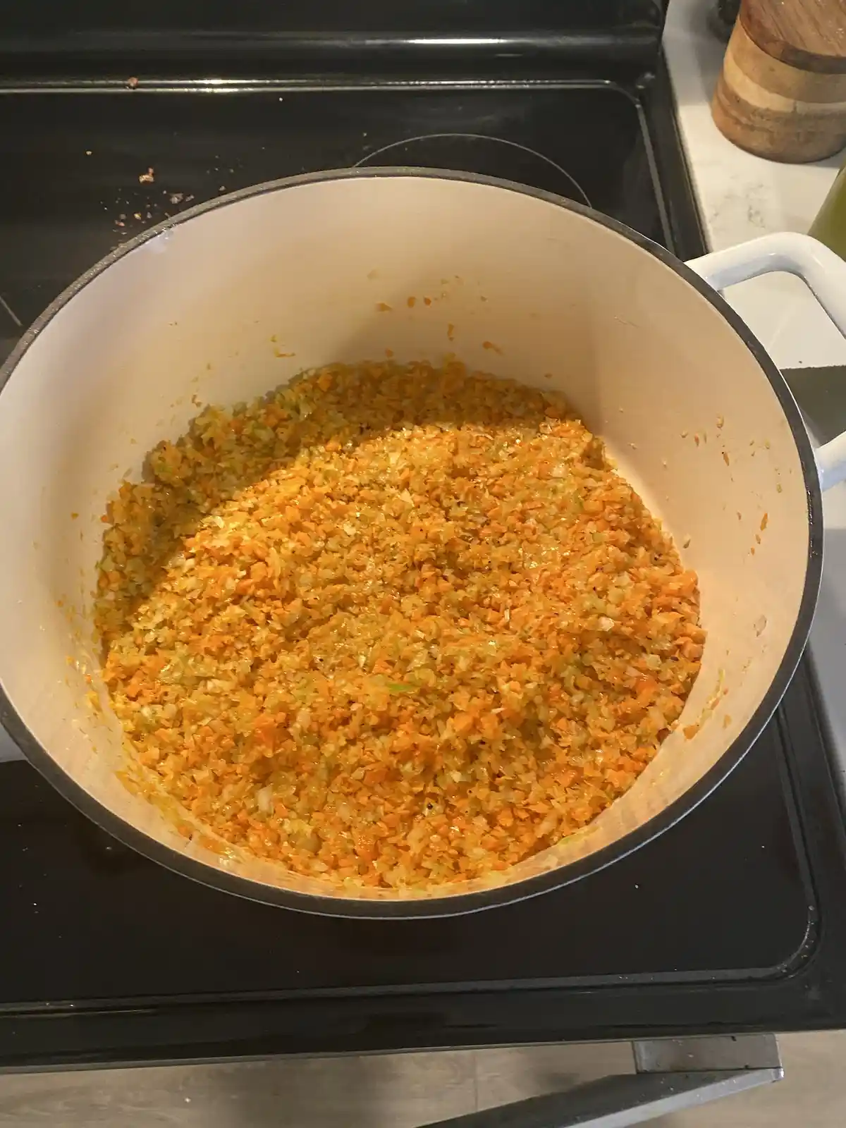 A large white enameled pot sits on a black stovetop, filled with a vibrant orange mixture of finely chopped carrots, onions…