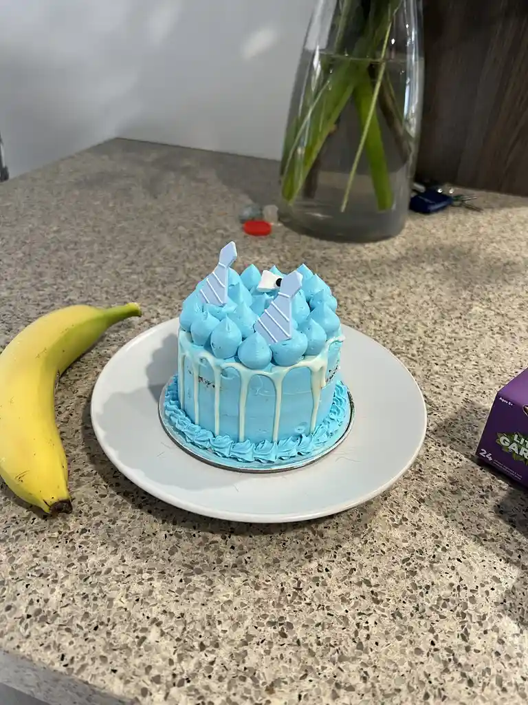 A small blue-frosted cake with white drip icing and two light blue tie decorations sits on a white plate on a speckled…