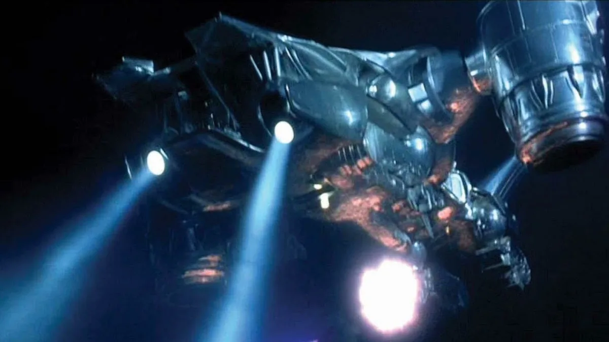 A large, damaged, metallic robot with glowing blue lights and exposed internal wiring is shown against a dark background…