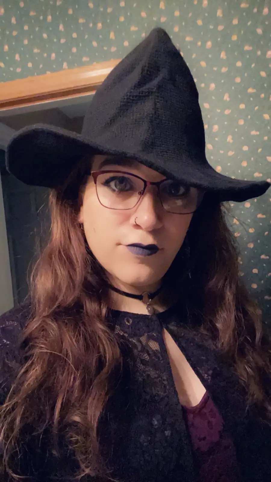 A person with long brown hair, wearing a black witch hat, glasses, dark lipstick, and a black lace top, stares directly at…