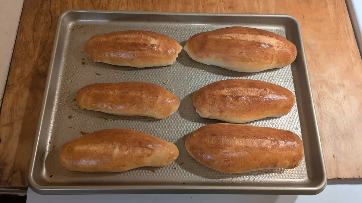 A metal baking sheet holds six golden-brown, oblong loaves of bread arranged in three rows on a wooden surface. The bread…