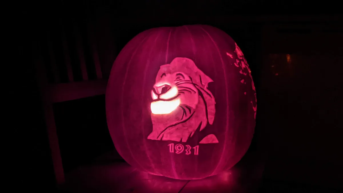 A carved pumpkin illuminated from within, casting a pink glow, features a detailed portrait of a smiling lion's head…