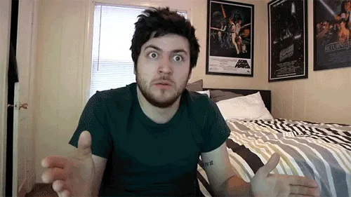 A man with a beard and dark hair sits on a bed with striped sheets and Star Wars posters on the wall behind him. He has a…