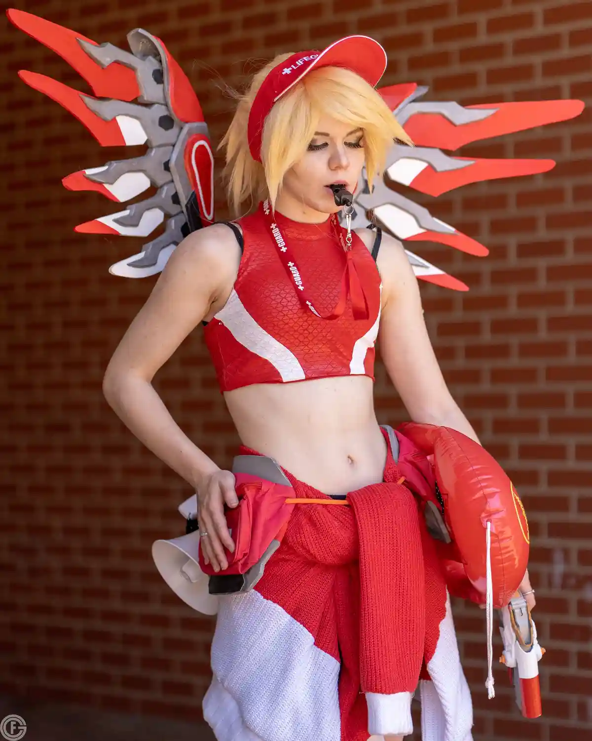 A person in a red and white lifeguard costume with large red and silver wings, holding a whistle to their mouth, a…