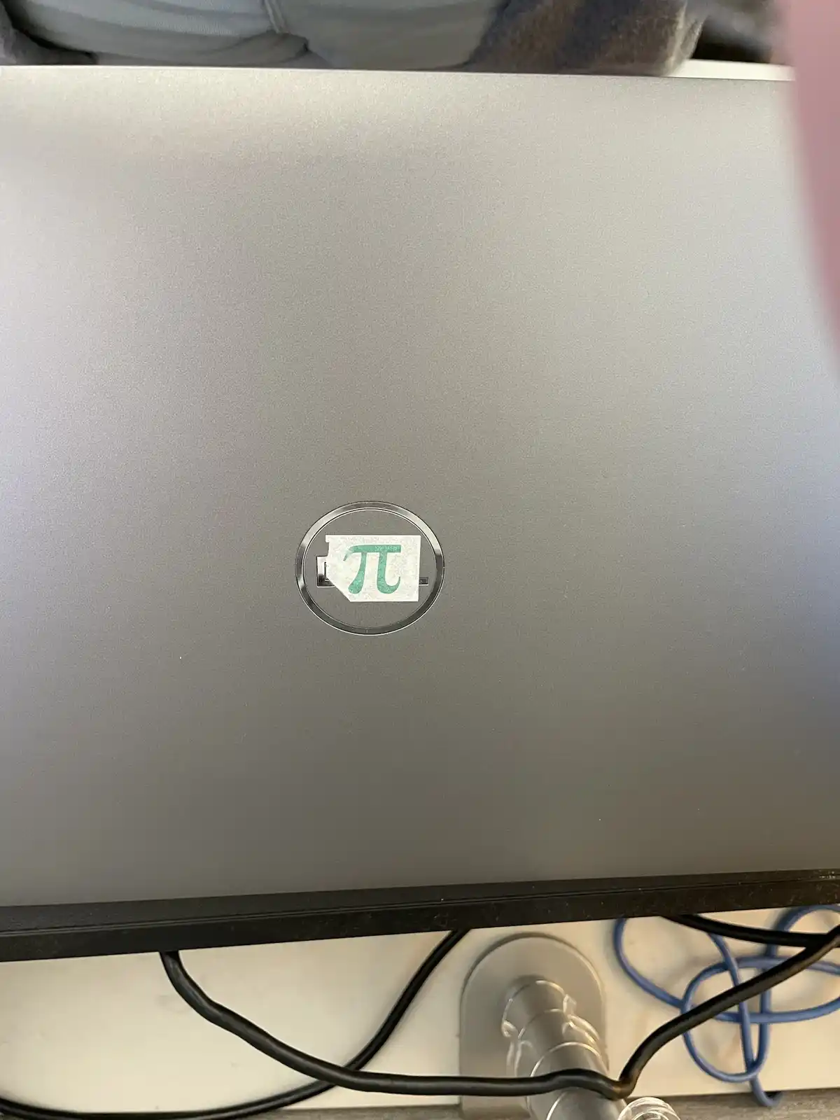 A close-up photo shows the lid of a silver laptop, where the standard Dell logo has been replaced by a green pi symbol (π)…