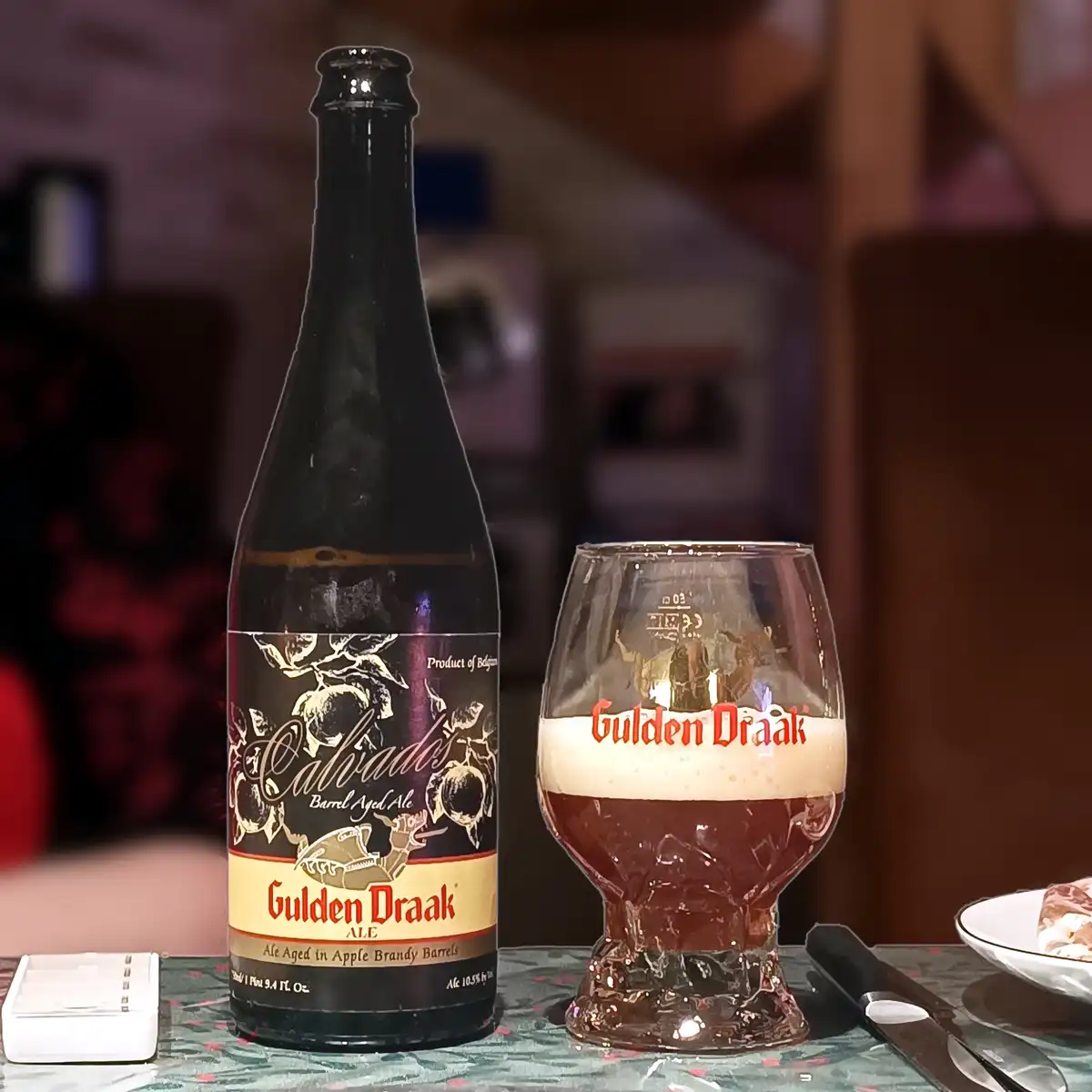 A dark bottle of Gulden Draak Cabriados Barrel Aged Ale sits next to a tulip glass filled with the same beer, which has a…