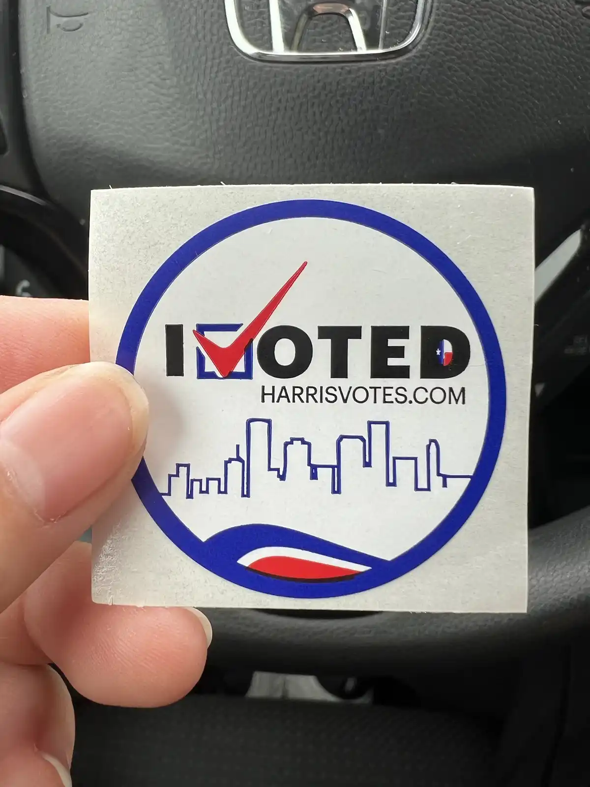 A hand holds a square 'I VOTED' sticker in front of a car's steering wheel. The sticker features a blue circle with a red…