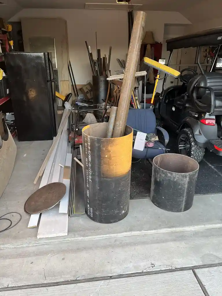 A cluttered garage contains a large metal pipe filled with long metal rods, next to an empty pipe, a blue chair, a golf…