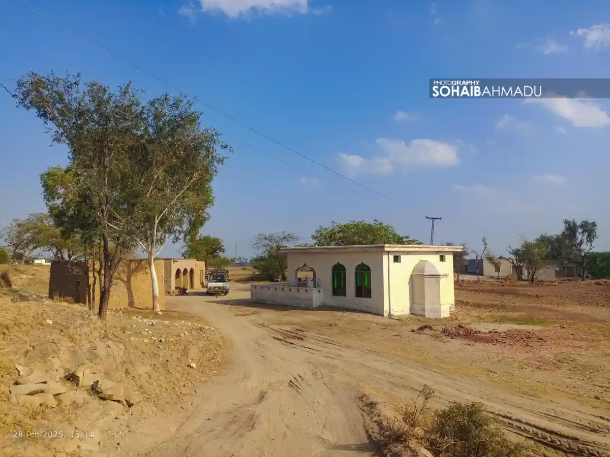 A rural dirt road scene under a clear blue sky, featuring a small, newly built white building with arched windows and a…