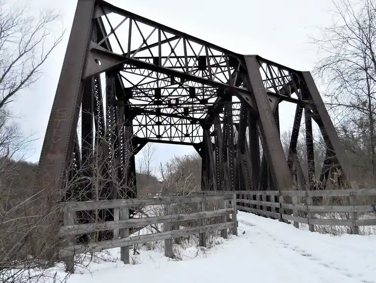 A low-angle photo of a rusty, snow-covered metal truss bridge with bare trees in the background. Graffiti reading 'STEVE ♥…
