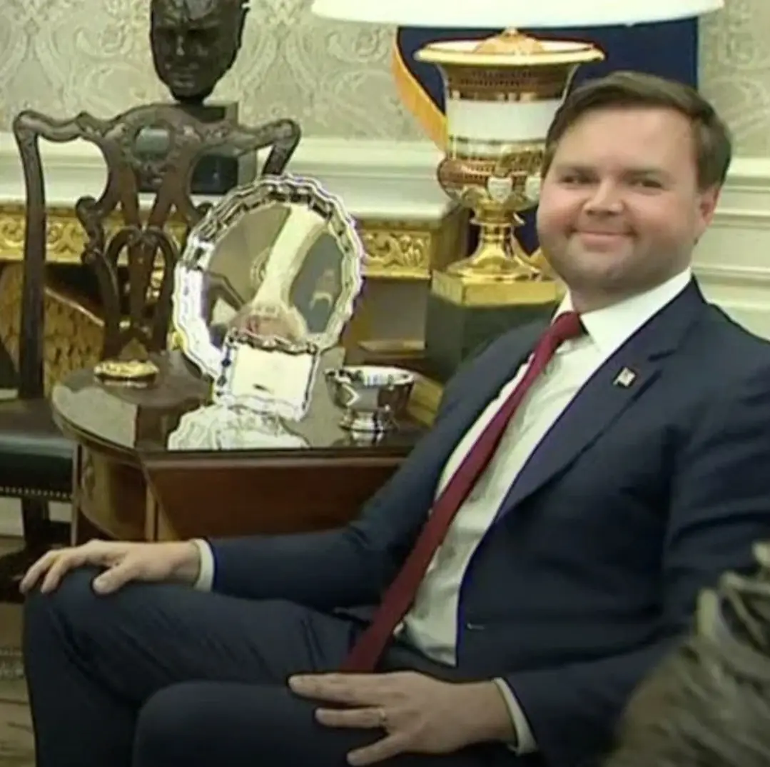 a man in a dark suit and red tie sits in an ornate room with gold-trimmed furniture, smiling slightly. Behind him, a large…