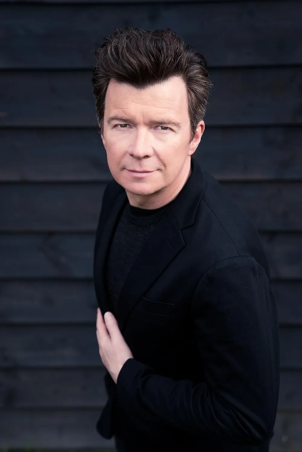 A man with dark hair and a black blazer stands against a dark wooden wall, looking directly at the camera with a subtle…