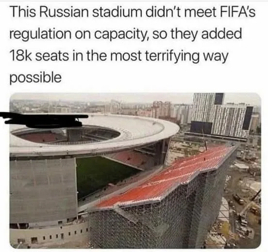 An aerial view of a large, modern stadium with a distinctive curved roof and a partially constructed, jagged, multi-tiered…