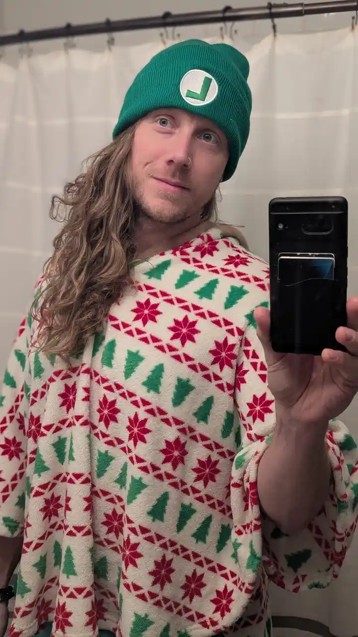 A person with long hair and a nose ring takes a mirror selfie in a bathroom, wearing a green Luigi beanie and a festive…
