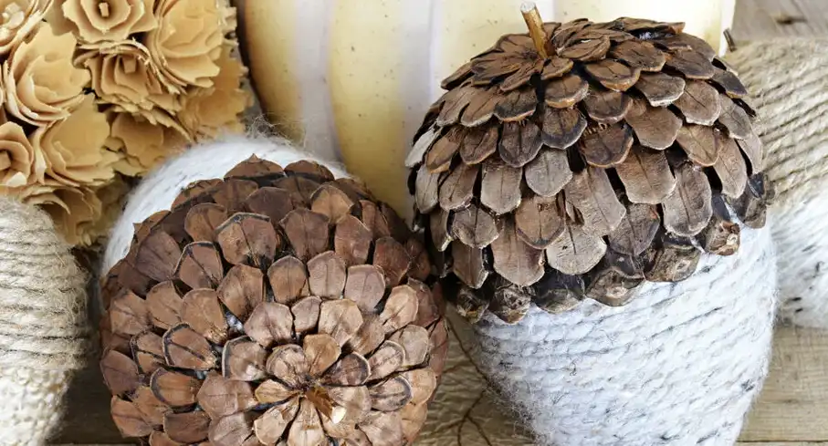Two pinecones are glued onto white yarn-wrapped bases to resemble acorns, surrounded by fall decor like paper flowers and…