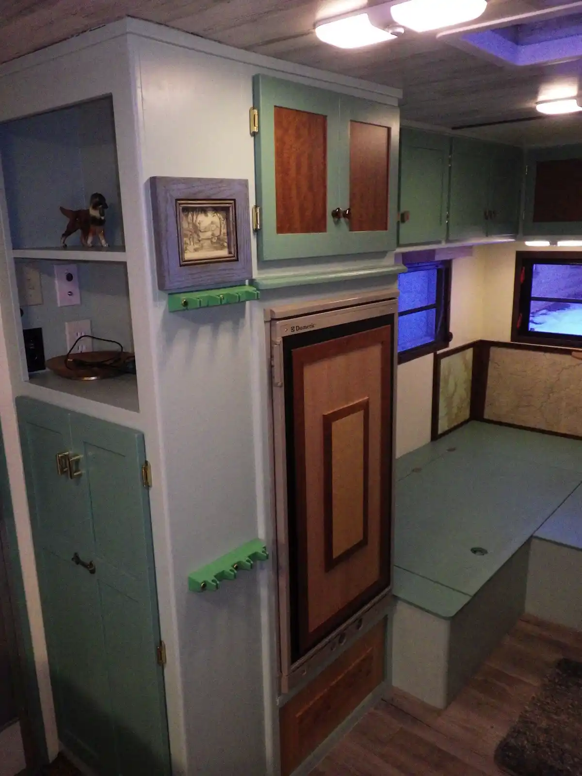 An interior shot of a compact, custom-built RV with light green cabinets, wooden accents, and a built-in bed. A Dometic…