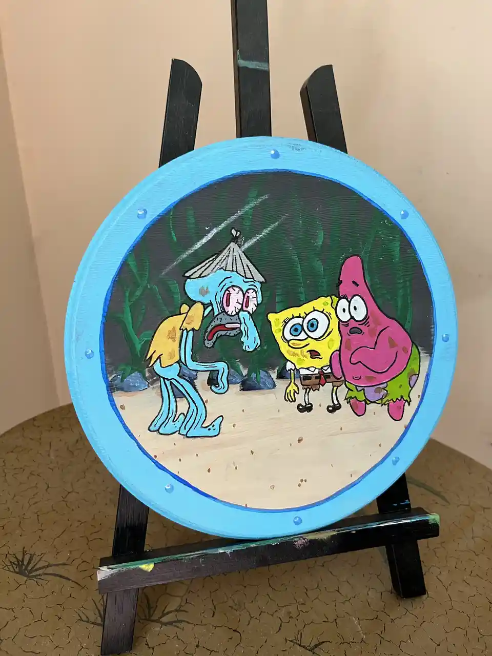 A hand-painted circular artwork on a blue-framed disc, displayed on a black easel, depicts three characters from SpongeBob…